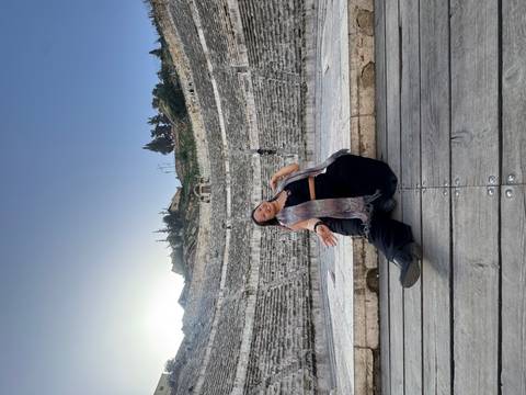 Traveller poses on wooden stage boards inside the ancient Roman amphitheatre with stepped stone seating.