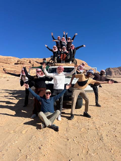 Cheerful group of travellers posing with arms raised on top of an open 4x4 jeep in the desert.
