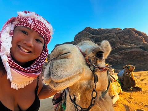 Close-up selfie of smiling traveller with friendly camel against rocky desert backdrop.