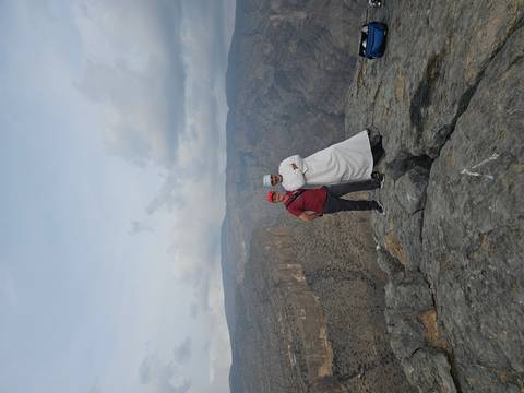 Two men stand on a rocky cliff edge overlooking a dramatic canyon landscape under a cloudy sky.