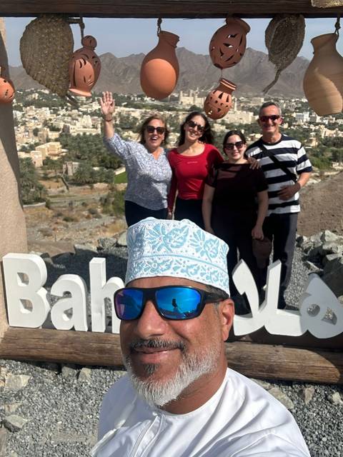 A cheerful group selfie in front of large white letters spelling Bahla with a desert valley backdrop.