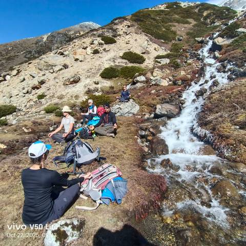 Trekkers sit meditating beside a cascading alpine stream amid rocky slopes.