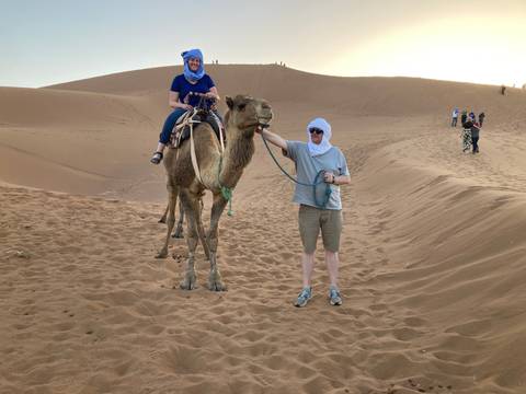 Traveller leads a camel with partner seated on its back amid rolling Sahara dunes at sunset.