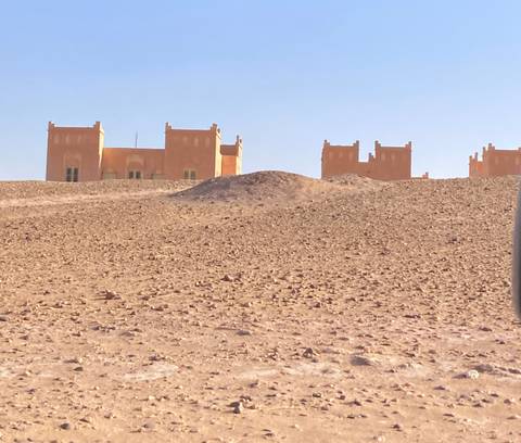 Isolated adobe-style buildings sit on a barren rocky desert plain beneath clear blue sky.