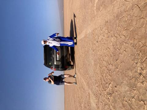 Tourist and local guide pose beside 4x4 vehicle on cracked desert lakebed under bright sky.