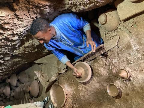 Man in blue robe shapes clay on a pottery wheel inside an earthen workshop.