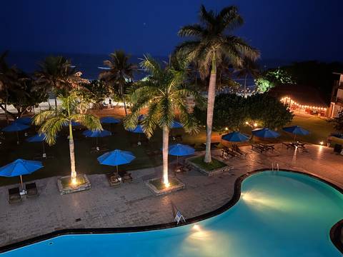 Nighttime view over illuminated resort pool, palm trees and beach with dark ocean beyond.