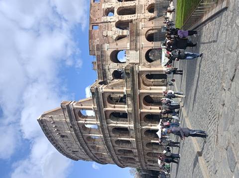 Street-level view of the Colosseum with many tourists walking on cobblestones under a blue sky with scattered clouds.