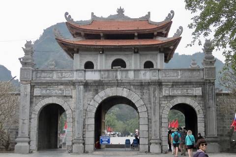 Stone entrance gate with three arches set against limestone peaks, visitors walking below.