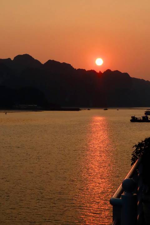 Orange sun setting behind dark karst cliffs reflecting light across a broad bay.