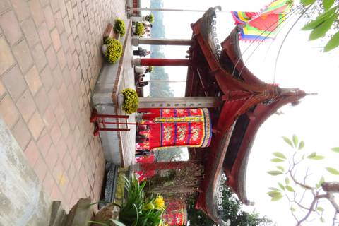 Open pavilion with red roof and decorated drum overlooking a lake, visitors gathered around.