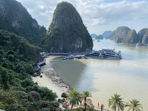 Panoramic view of a small sandy cove and pier surrounded by the limestone karst islands of Halong Bay with several cruise boats docked.