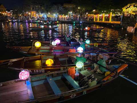 Colorful lantern-lit wooden boats on a river at night in front of a lively waterfront packed with people.