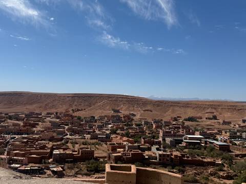 Brown adobe town sprawls across a desert ridge with high plateau and snowy peaks in background.