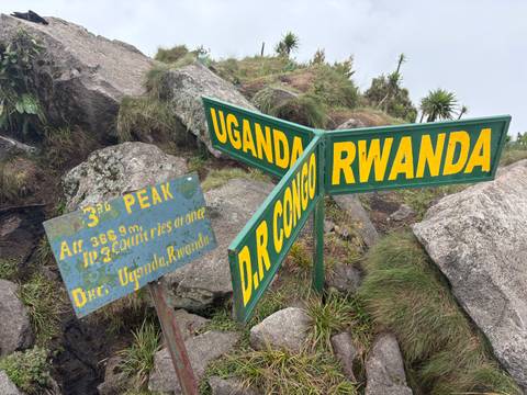 Trail summit sign marking the borders of Uganda, Rwanda and D.R. Congo amid misty rocks.