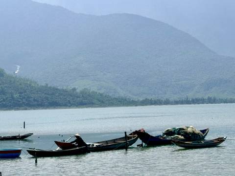 Small wooden fishing boats float on a calm lagoon backed by hazy mountains.