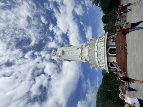 Towering white Lady Buddha statue rises against a dramatic cloud-filled sky.