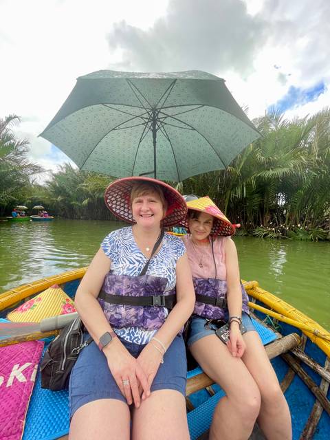 Two smiling women wearing conical hats sit in a round bamboo boat on a green river, shaded by an umbrella.