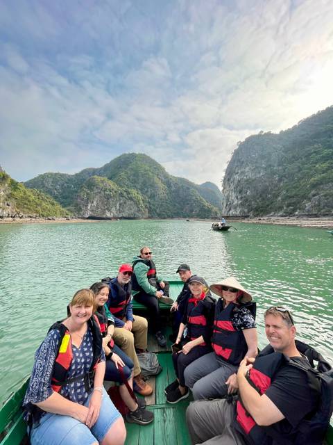 Travelers in life-jackets sit in a small boat surrounded by green karst cliffs and calm emerald water.