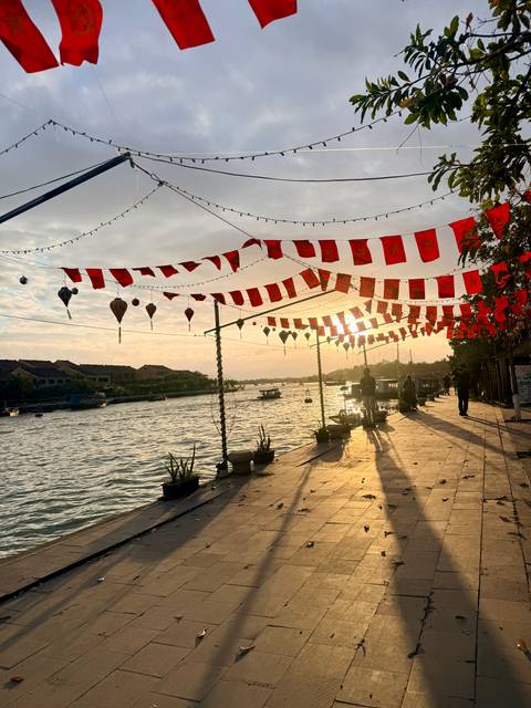Riverside promenade decorated with red lanterns and flags at sunset, casting long shadows on the pavement.