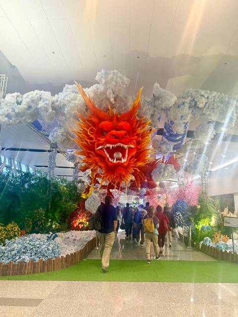Indoor walkway filled with travelers beneath a dramatic red dragon decoration suspended among artificial clouds.