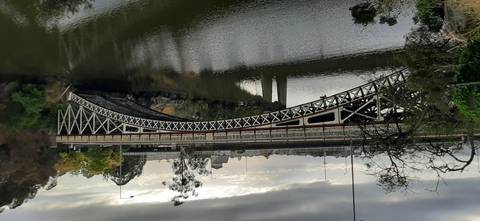 Ornate iron bridge spans a calm river beneath grey skies, reflections rippling in the water.
