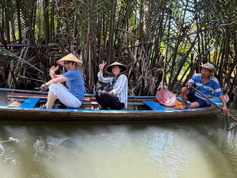 Small wooden sampan gliding through dense palms with smiling passengers in conical hats.