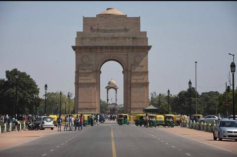 Busy roadway leads to the grand sandstone India Gate monument under a hazy sky