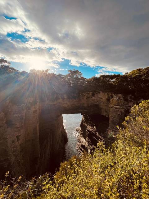 Sunbeams stream over a natural sea arch carved into rugged cliffs above the ocean.