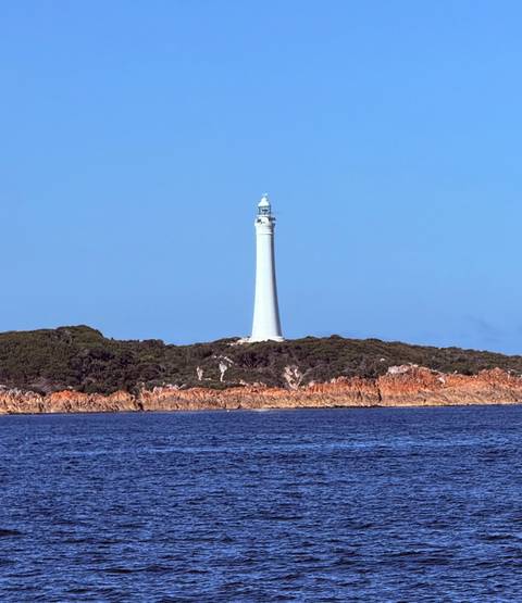 A solitary white lighthouse rises above a rocky, scrub-covered island against a clear sky.