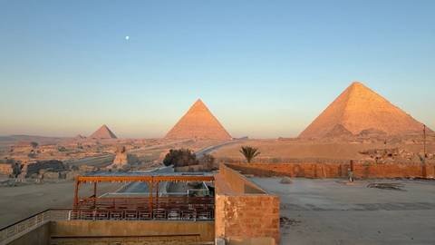 Panoramic sunrise view of the Pyramids of Giza with soft golden light and a faint moon overhead.