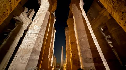 Night view looking upward between towering hieroglyph-covered columns toward an obelisk at Luxor Temple.