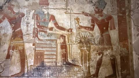 Ancient Egyptian wall relief with seated pharaoh receiving an offering, faded pigments on stone inside a temple.