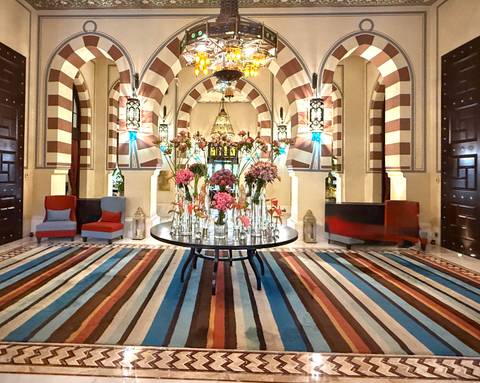 Elegant hotel lobby with striped Moorish arches, chandelier and colorful flower arrangement on a round table.