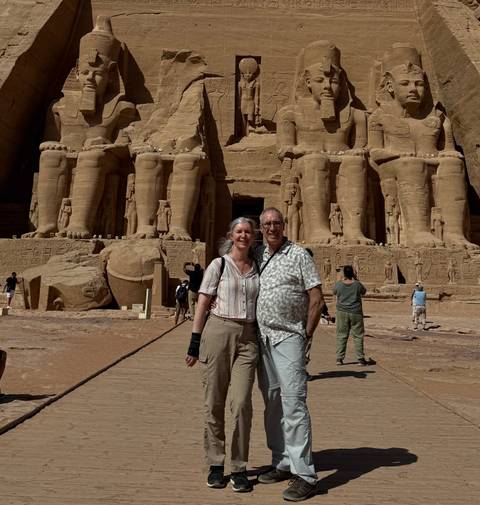 Smiling couple posing in front of the colossal statues of Abu Simbel temple under bright sun.