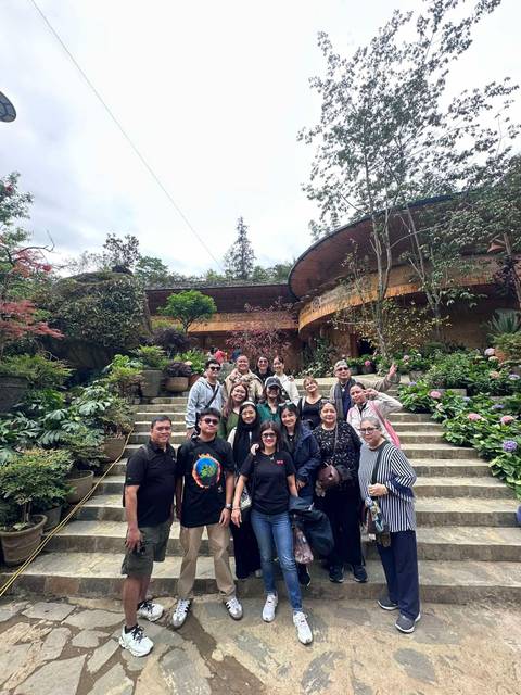Large tour group poses on stone steps surrounded by lush gardens and flowers at a hilltop attraction.