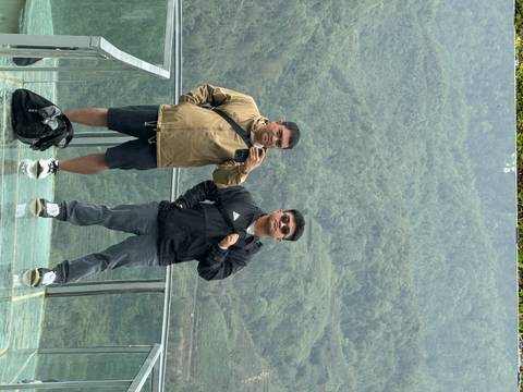 Two travelers pose on a glass skywalk overlooking steep green valleys and terraced hills.