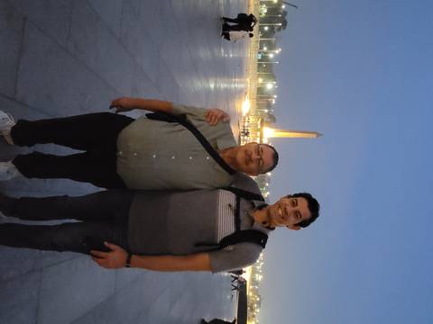 Two travelers arm-in-arm posing at dusk in a large square with illuminated obelisk in background.