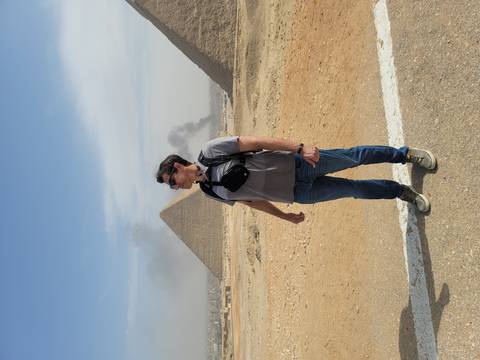 Man standing in front of the Great Pyramid with plumes of smoke in distant sky.