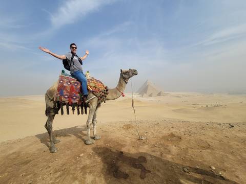 Traveler riding a decorated camel in the desert with the Pyramids of Giza rising through light haze behind.