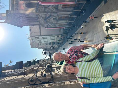 Couple poses on a balcony overlooking a pedestrian street lined with historic buildings under bright sunlight.