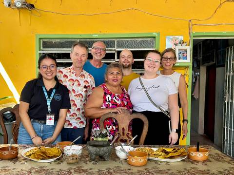 Group of travelers and locals smiling around a table after sharing a home-cooked meal in a brightly painted room.