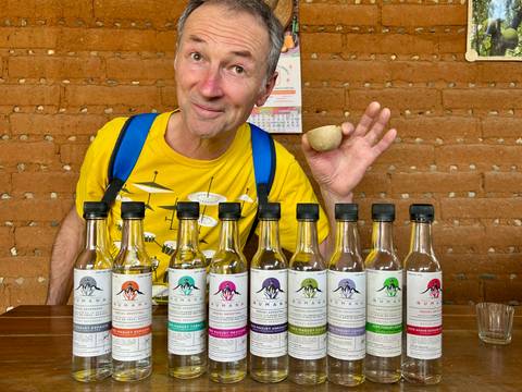 Man proudly displays a colorful lineup of artisanal mezcal bottles against an adobe wall.