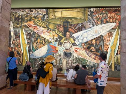 Visitors observe and photograph a vibrant Diego Rivera mural inside a museum hall.