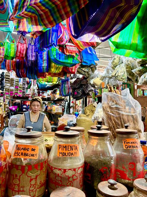 Market vendor surrounded by colorful hanging textiles and piles of spices inside a bustling bazaar.