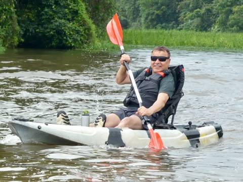 Smiling man paddles a white kayak across calm brown river water, splashing with each stroke.