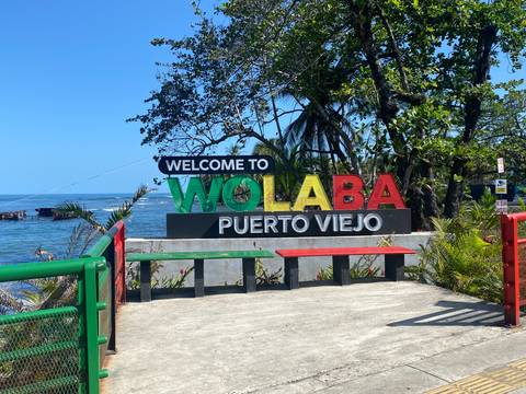 Colorful coastal sign reading 'Welcome to Wolaba Puerto Viejo' overlooks blue Caribbean waters and palm trees.