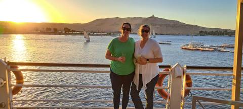 Two women enjoy drinks on a Nile cruise deck at golden sunset with feluccas sailing past sandy Aswan hills.