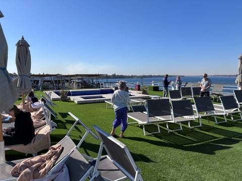 Passengers lounge on a sunny Nile cruise sundeck with pool, deckchairs, and river views.
