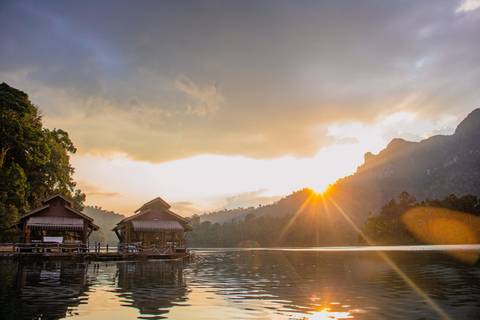 Floating raft houses rest on a glassy lake in Khao Sok National Park as the rising sun casts rays over misty jungle-covered mountains.